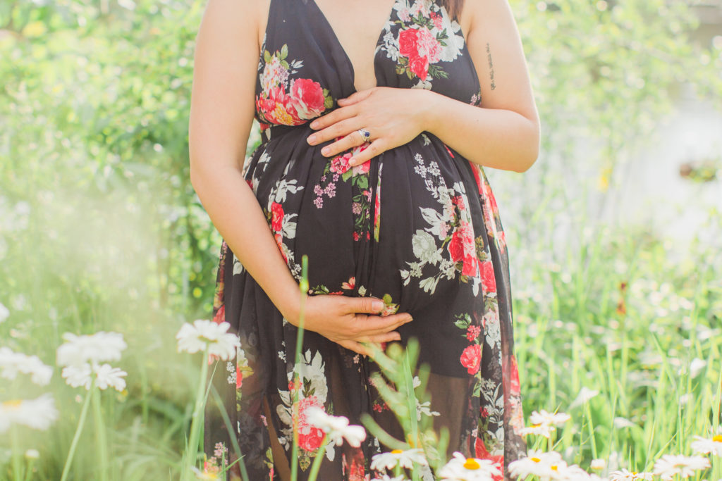 Maternity close up belly photo. Mom is holding her baby belly standing in-between tall grass and flowers