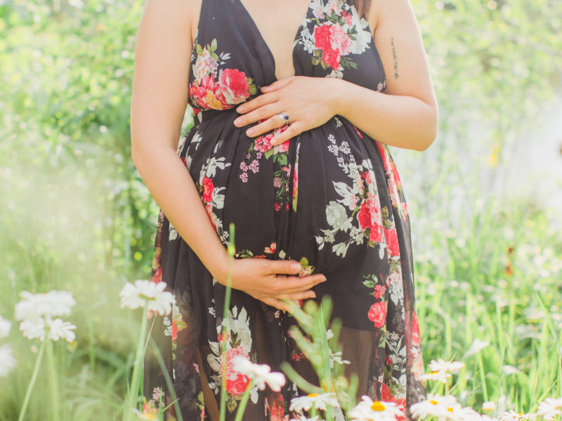 Maternity close up belly photo. Mom is holding her baby belly standing in-between tall grass and flowers