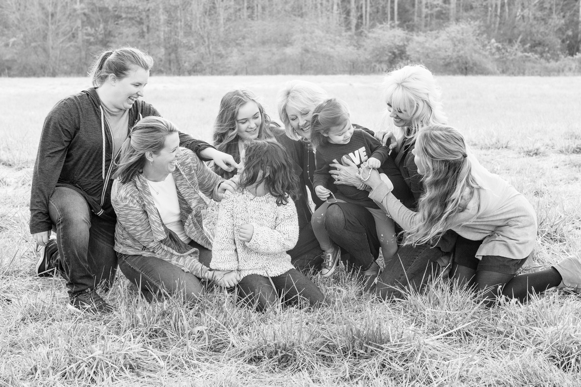 Photo of all the woman during an extended family session. Woman are kneeling in the grass laughing at each other