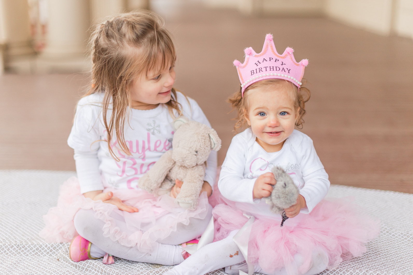 Sisters sitting next to each other during first birthday session. Older sister is looking down on one year old, smiling at her.