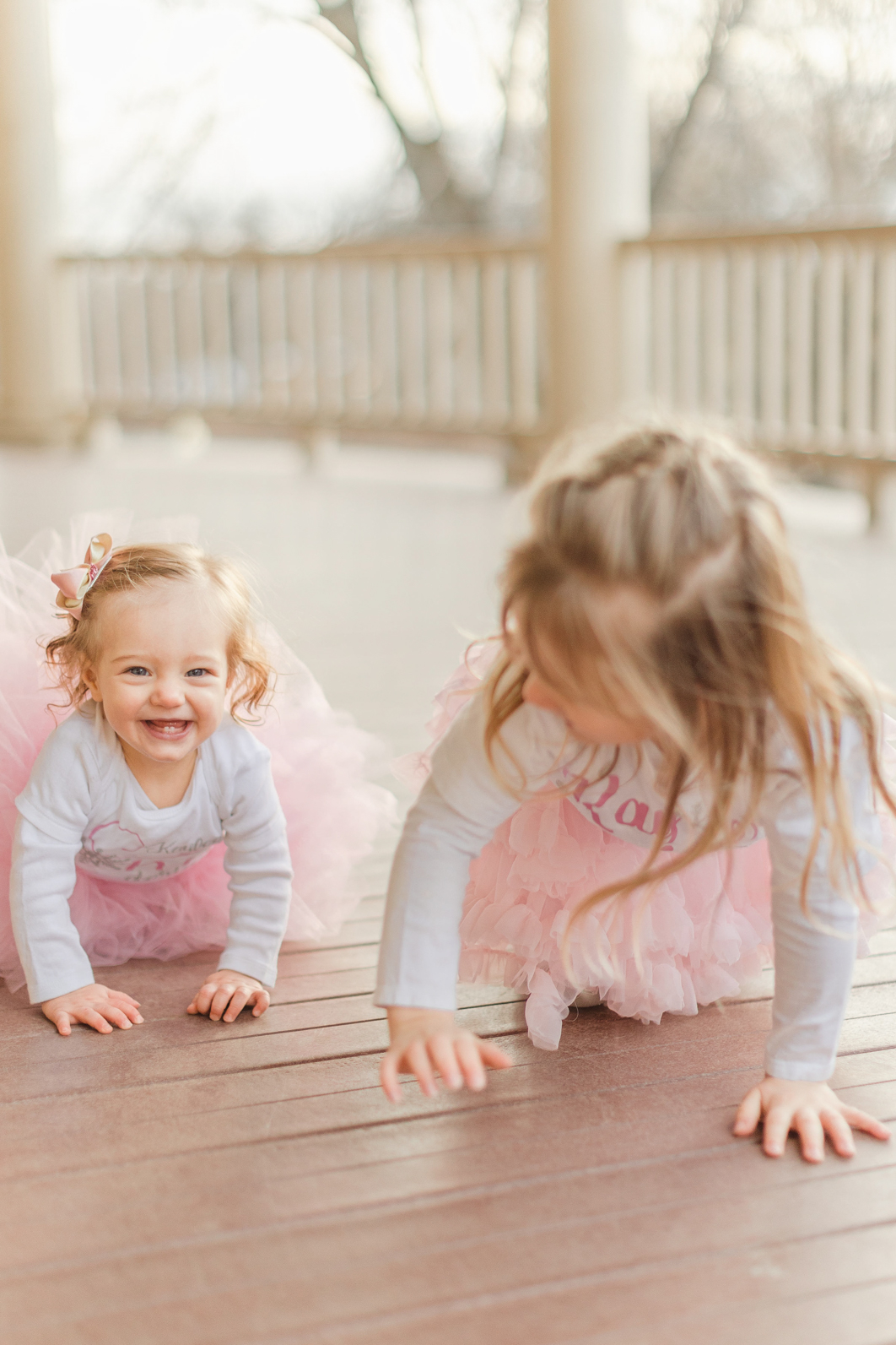 Sisters are crawling towards the camera laughing. Both are wearing a white shirt and a pink tutu