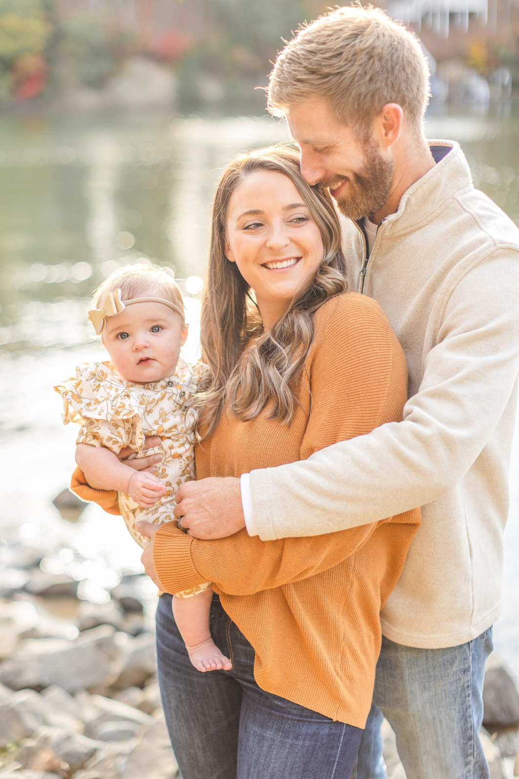 Fall Family mini session in Charlotte NC. Dad is hugging mom from behind. Mom is holding daughter. Mom is smiling onto the distance over her shoulder. Dad is nuzzling moms temple.