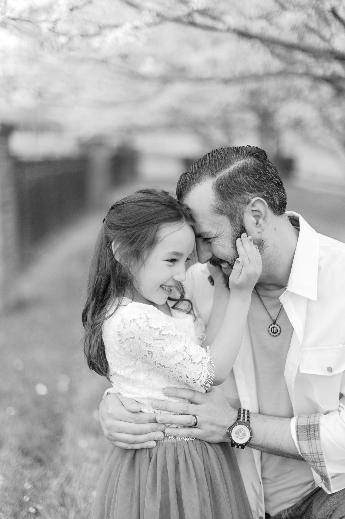 Black and white photo of dad kneeling next to his daughter, who is cuddling and laughing with him, while she is holding his cheeks during their charlotte spring minis