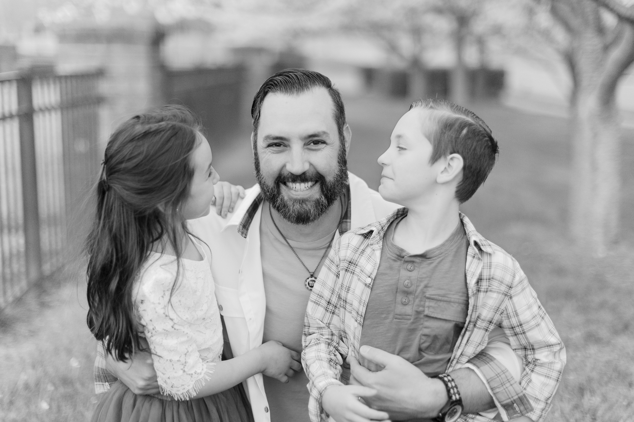 Black and white photo of dad kneeling down, having both, his daughter and his son, on his knees. Kids are looking at their dad smiling, while dad is smiling into the camera. This was captured during their Charlotte Spring Minis