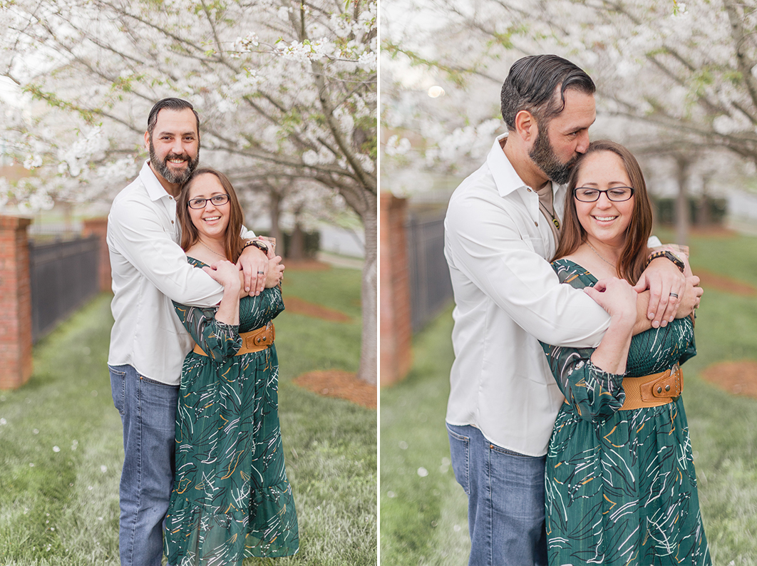 Mom and Dad are standing in front of a white cherry blossom tree. Dad is hugging mom from behind and giving her a kiss on the head, while she smiles down, looking over her shoulder during their charlotte spring minis