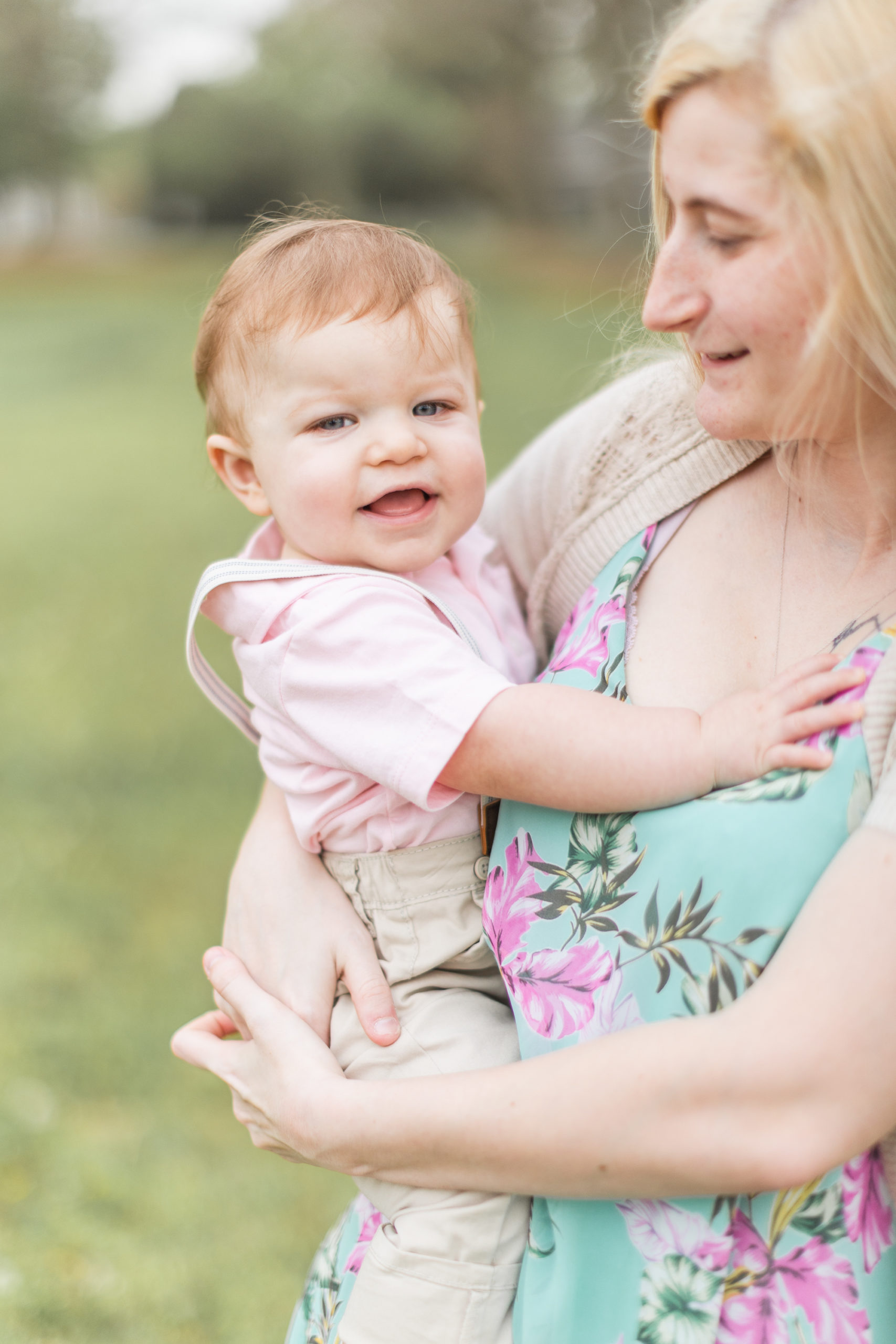 Mom is carrying her one year old son, smiling at him, while le laughs at the camera during their mommy + me cake smash session