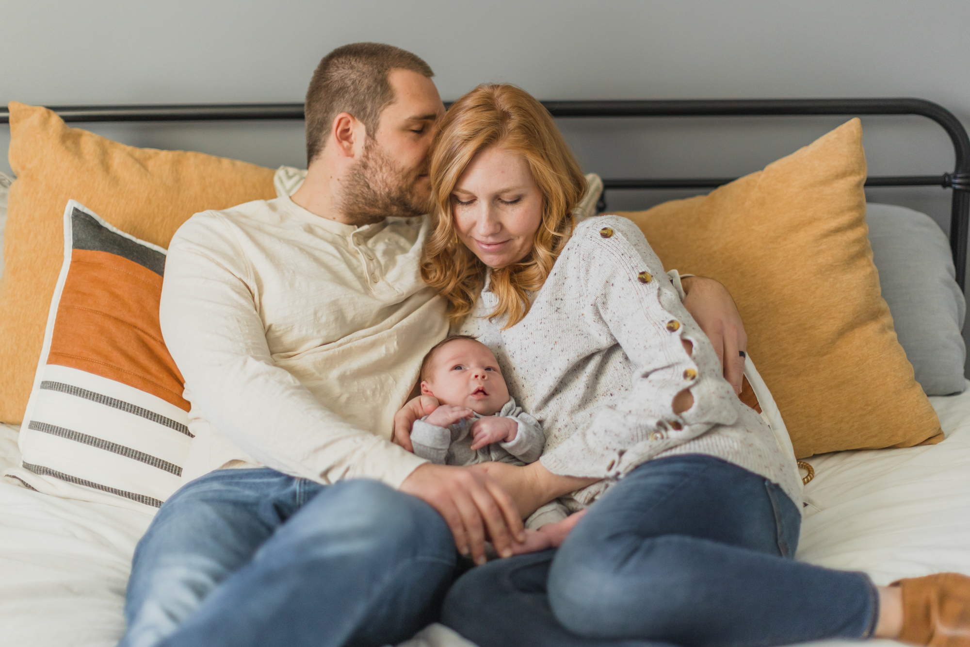 Mom and Dad are sitting on their bed next to each other, while holding their newborn in their arms during their in-home lifestyle newborn session