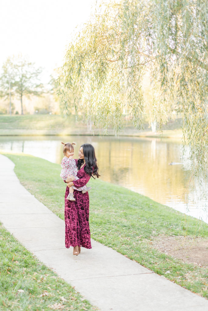 Mother and Daughter walking towards the camera. Mom is holding her daughter and smiling at her. They are next to a willow tree on a walking path by a pond during sunset